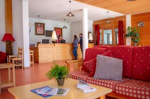 A hotel lobby with a reception desk, wooden furniture, a red patterned sofa, and a plant on a table.