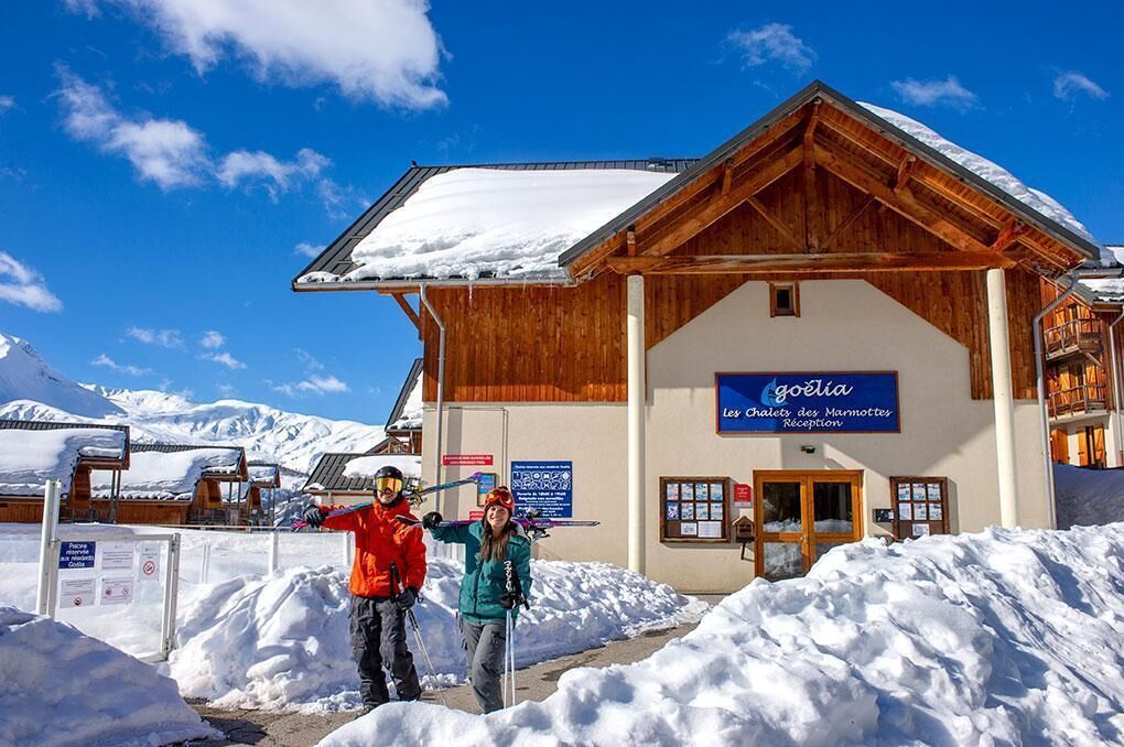 Un bâtiment en bois avec une enseigne indiquant « Goélia Les Chalets des Marmottes, réception », le tout sur fond de montagnes enneigées.