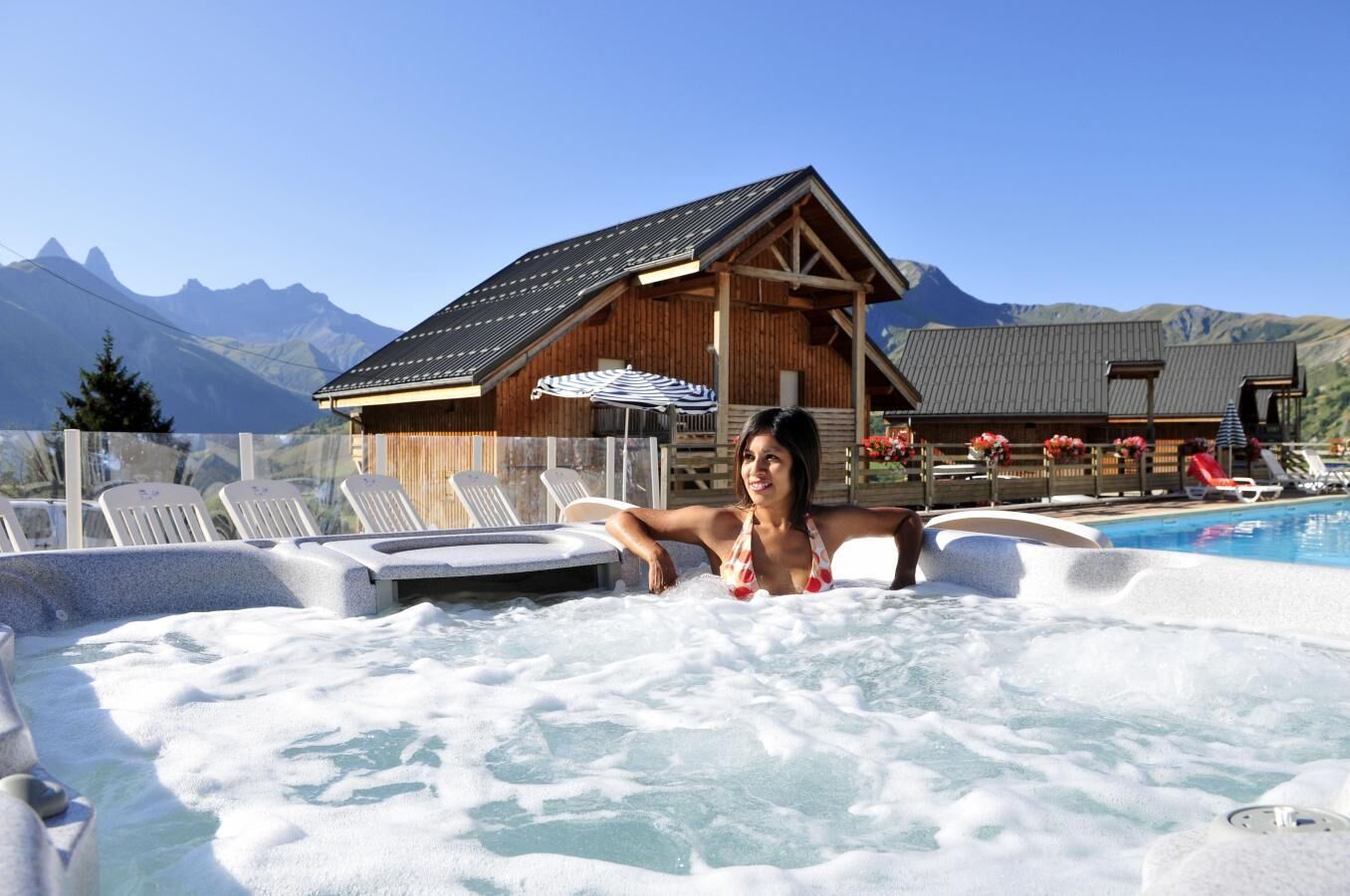 En person i ett bubbelbad med berg i bakgrunden och en träbyggnad med ett sluttande tak.