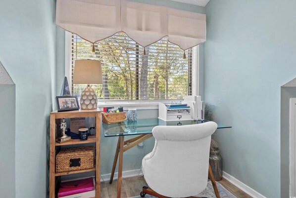 A study with a glass desk, a white chair, a wooden shelf with various items, and a window with a view of trees.