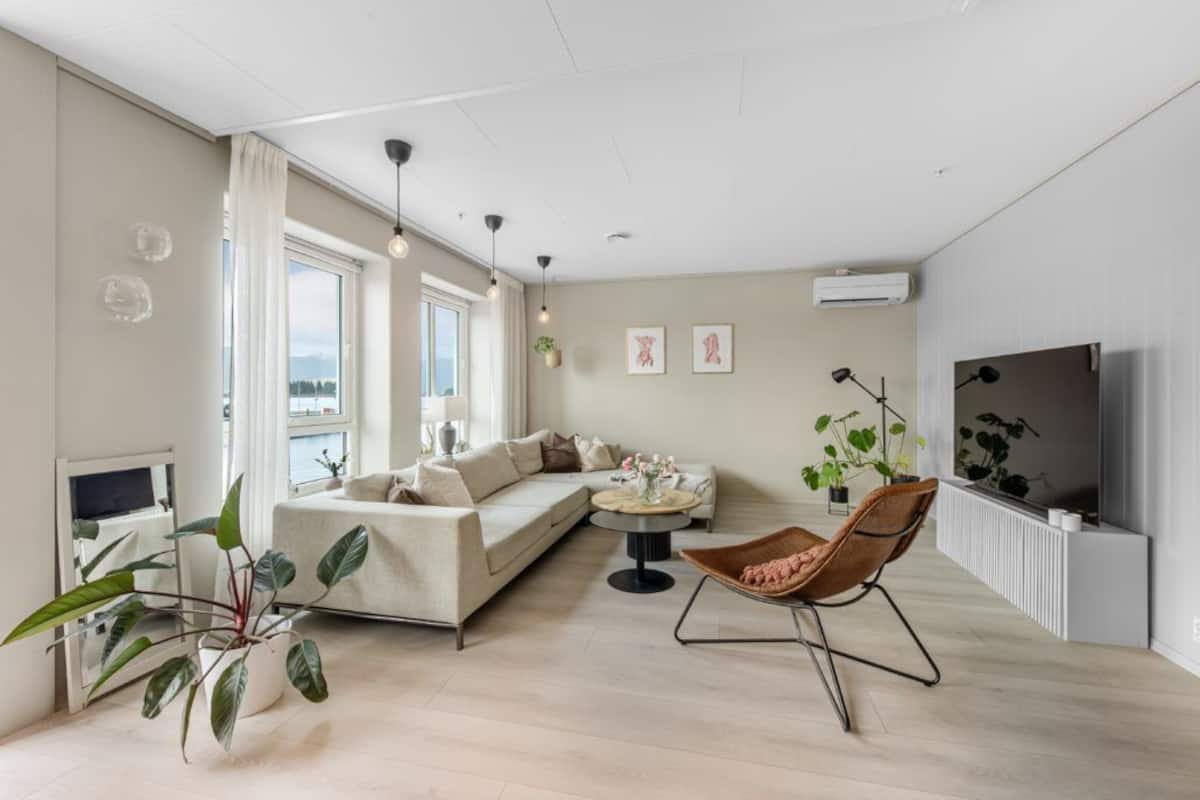 A modern living room with a sofa, a round coffee table, and a TV. There are plants and a window with curtains.
