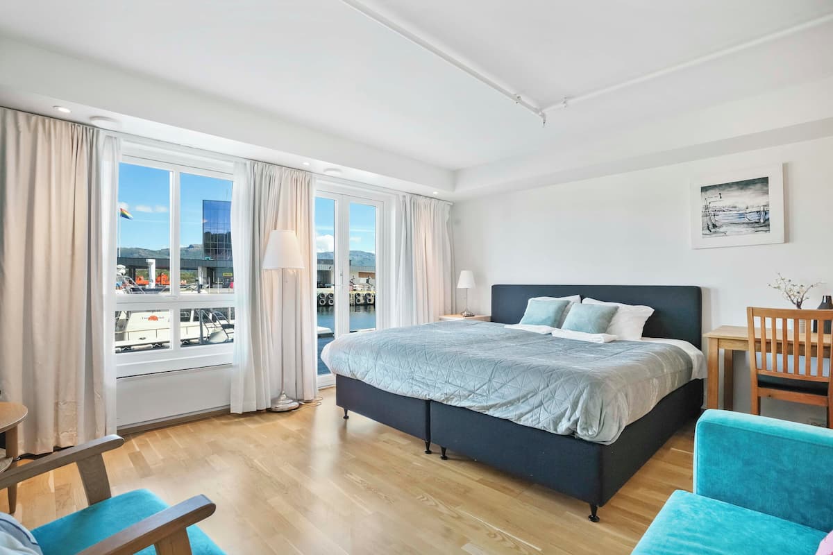 A modern hotel room with a large bed, a desk, and a view of the city through large windows.