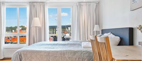 A hotel room with a bed, a wooden table, and a view of the outside through large windows.