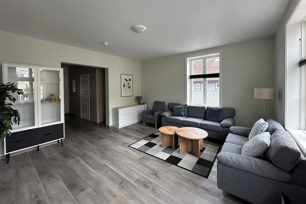 A modern living room with a grey sofa, a coffee table, and a cabinet. There is a window with blinds, a lamp, and a plant.