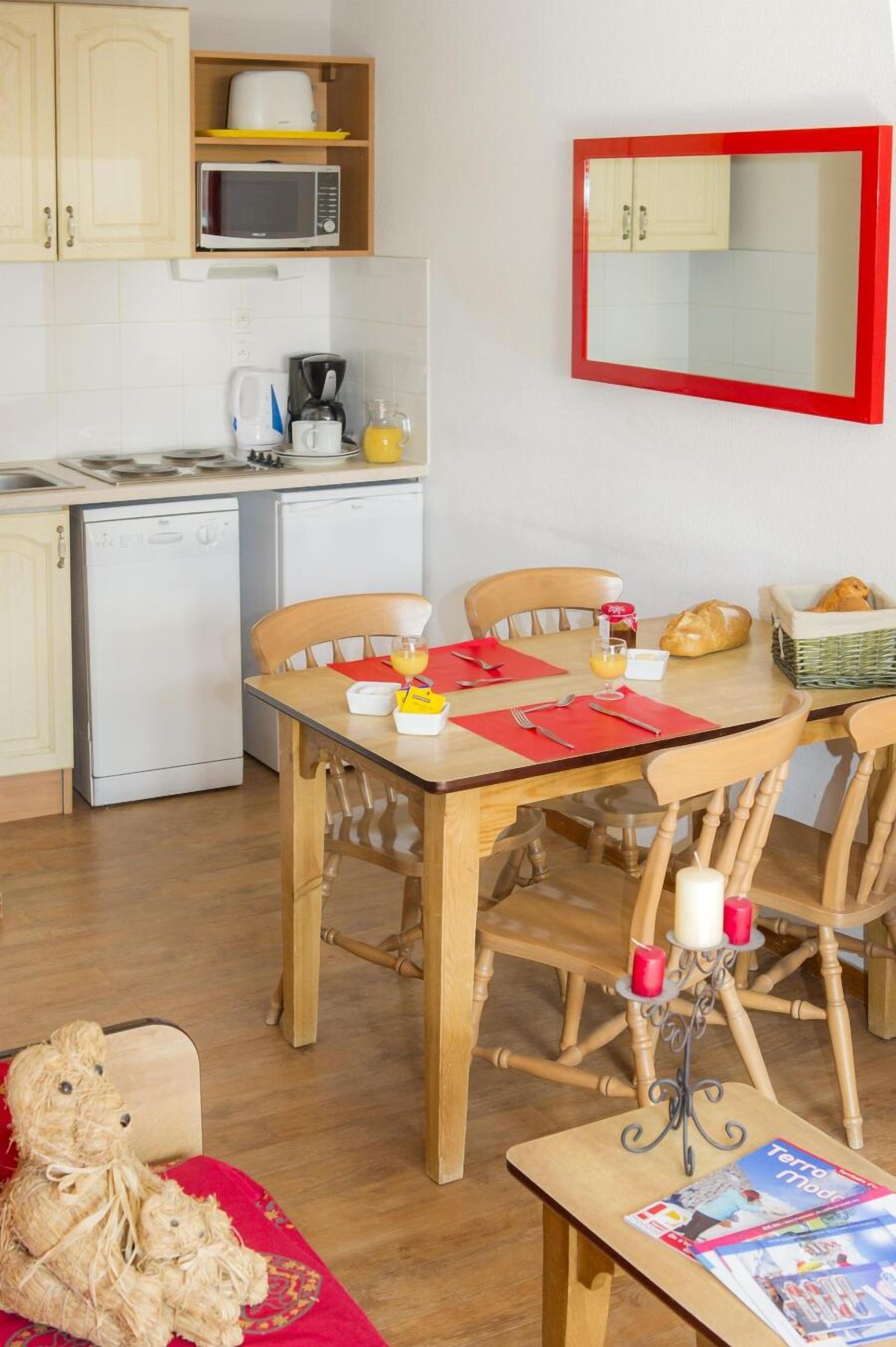 A compact kitchen with a microwave, coffee maker, and toaster on the countertop. There is a dining table with chairs, a small table with a candle, and a basket of bread.