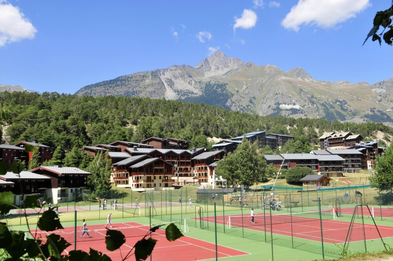 Un complex turístic a la muntanya amb diversos edificis d'estil xalet, una pista de tennis i un fons d'arbres.