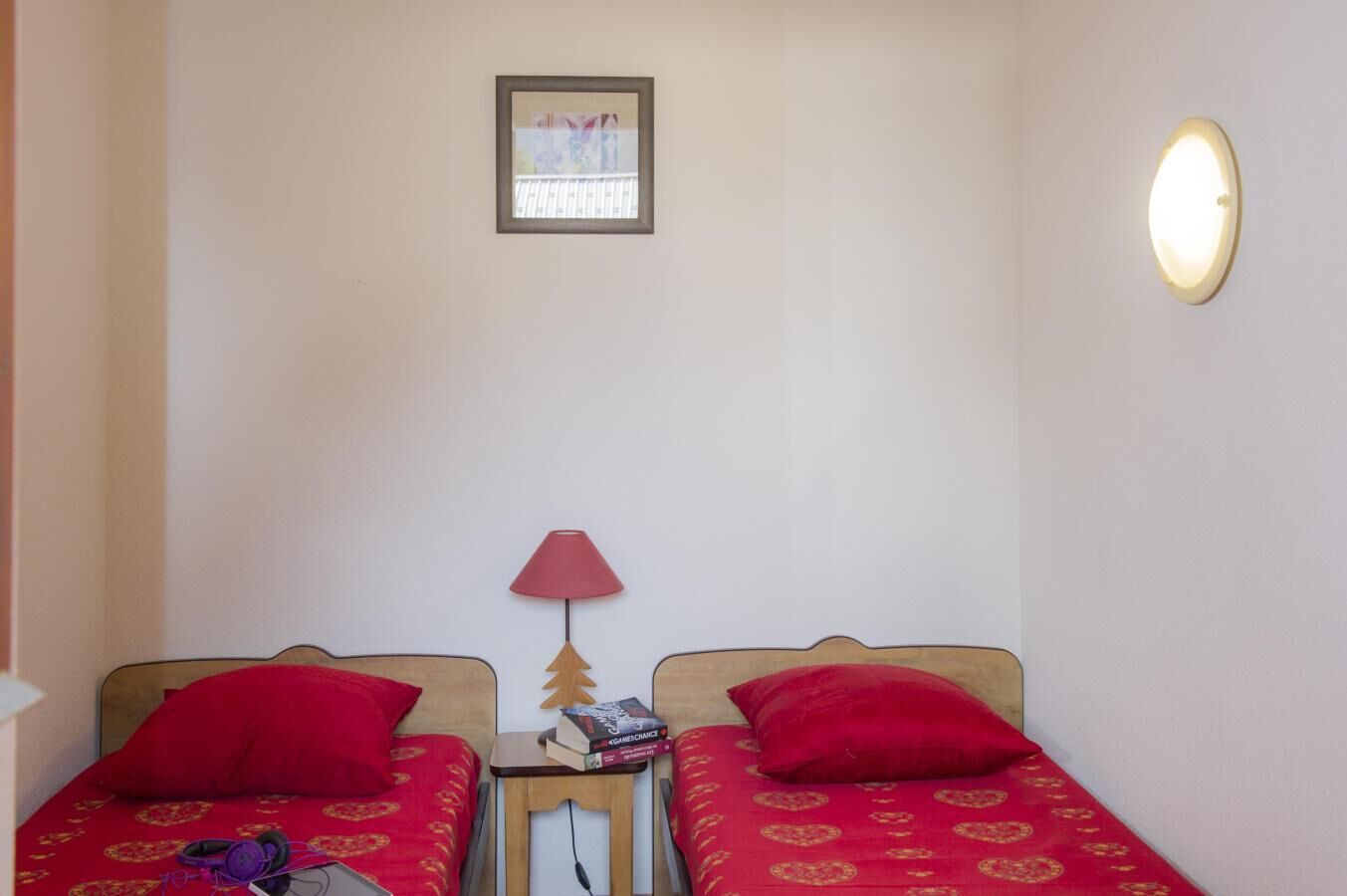 A small room with two single beds, each with a red bedspread and matching pillows. There is a small table with a lamp between the beds, and a framed picture on the wall above the beds.