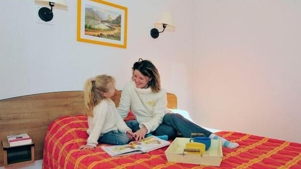 Une chambre avec un couvre-lit matelassé rouge et jaune, une table de chevet sur laquelle se trouvent un livre et une petite boîte, et une lampe fixée au mur.