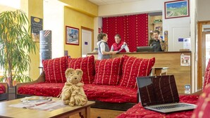 Et hotellobby med reception, en sofa med røde puder, en bamse på et bord og en bærbar computer på en seng.