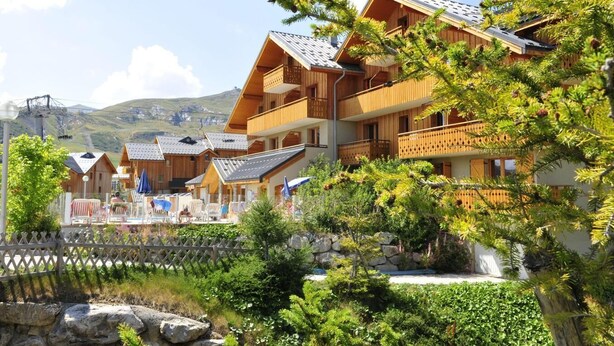 Et træchalet-lignende hotel med flere balkoner, omgivet af frodig vegetation og et bjerglandskab.
