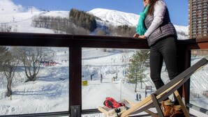A person standing by a window overlooking a snowy ski slope with a chairlift and buildings in the background. - 200m from ski slopes, fitness, swimming pool, sauna, terrace, balcony, tv, ski locker, 35-40m² (Le Corbier)