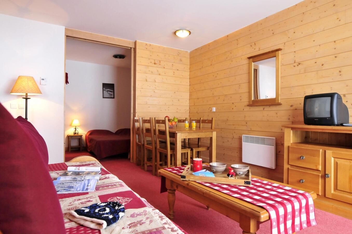 A cozy room with a wooden interior, a dining table with chairs, a bench, a television on a wooden stand, and a bed with a red blanket.