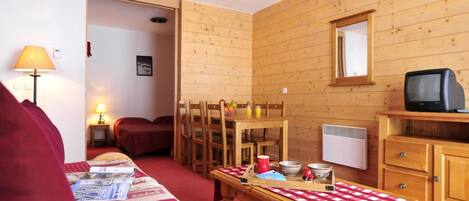 A cozy room with a wooden interior, a dining table with chairs, a bench, a television on a wooden stand, and a bed with a red blanket.