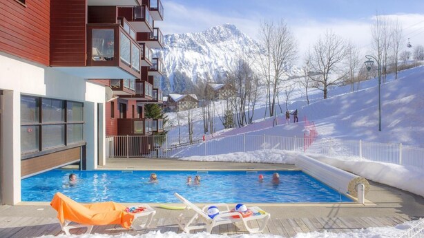 A swimming pool with lounge chairs, a building with balconies, and snow-covered mountains in the background. - 200m from ski slopes, fitness, swimming pool, sauna, terrace, balcony, tv, ski locker, 35-40m² (Le Corbier)