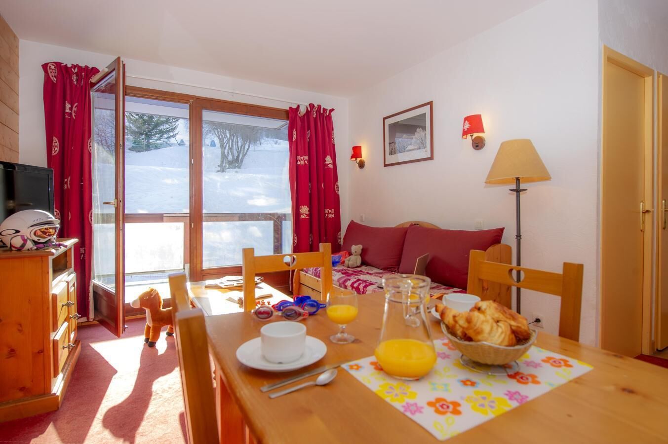 Une pièce chaleureuse avec une table à manger en bois, dressée pour le petit-déjeuner, un canapé avec des coussins rouges et une fenêtre ornée de rideaux rouges.