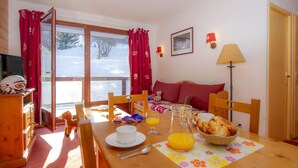 A cozy room with a wooden dining table set for breakfast, a sofa with red cushions, and a window with red curtains. - 200m from ski slopes, fitness, swimming pool, sauna, terrace, balcony, tv, ski locker, 35-40m² (Le Corbier)