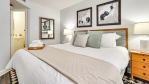 Un lit bien fait, recouvert d’une couverture beige, avec deux lampes de chevet et une table de nuit en bois sur laquelle se trouve une lampe. Des tableaux encadrés sont accrochés au mur au-dessus du lit.