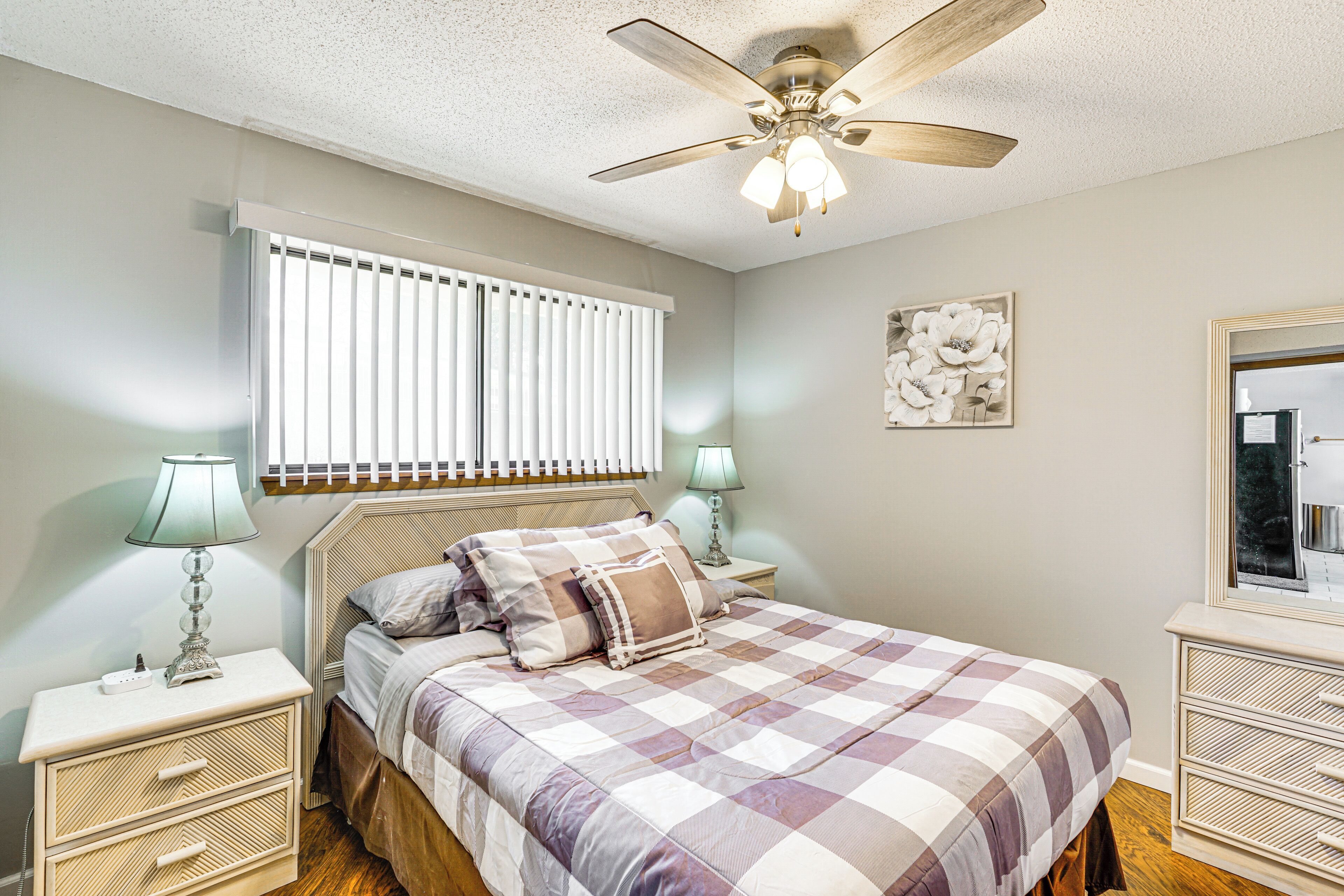 A bedroom with a bed, nightstands, a ceiling fan, a window with blinds, and a wall-mounted mirror.