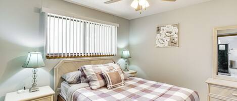 A bedroom with a bed, nightstands, a ceiling fan, a window with blinds, and a wall-mounted mirror.