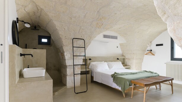 A bedroom with a stone ceiling, a bed with white linens, a bedside table, a wooden bench, and a small window.