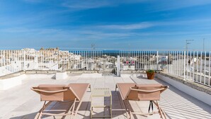 A rooftop terrace with white railings, two beige lounge chairs, and a potted plant.