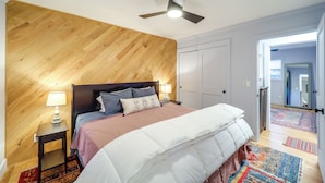 Un dormitorio con una pared de acento de madera, una cama con colcha blanca, dos lámparas de noche y un ventilador de techo.