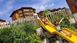 Um escorrega amarelo em área de jardim, com cercas de madeira e edifícios ao fundo.