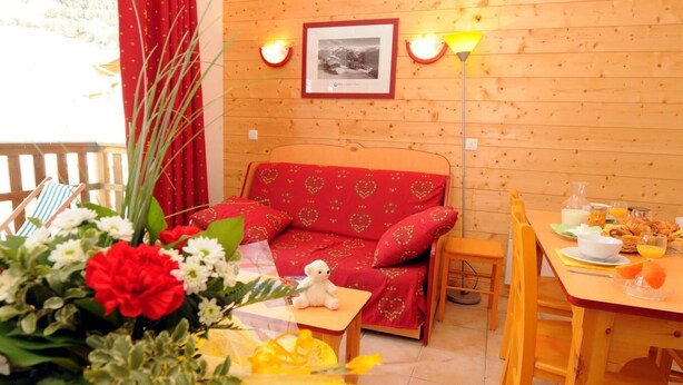 Une pièce chaleureuse avec un mur en bois, un canapé rouge orné de motifs en forme de cœur, une petite table avec des éléments pour le petit-déjeuner et un tableau encadré au-dessus du canapé.