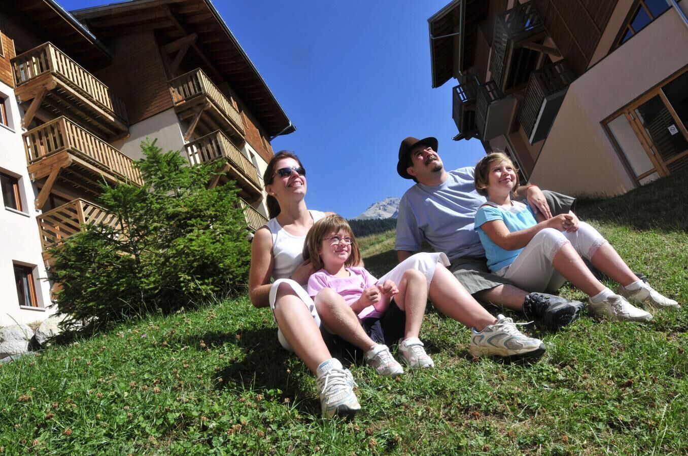 A group of people sitting on grass in front of a building with wooden balconies and a mountain in the background.