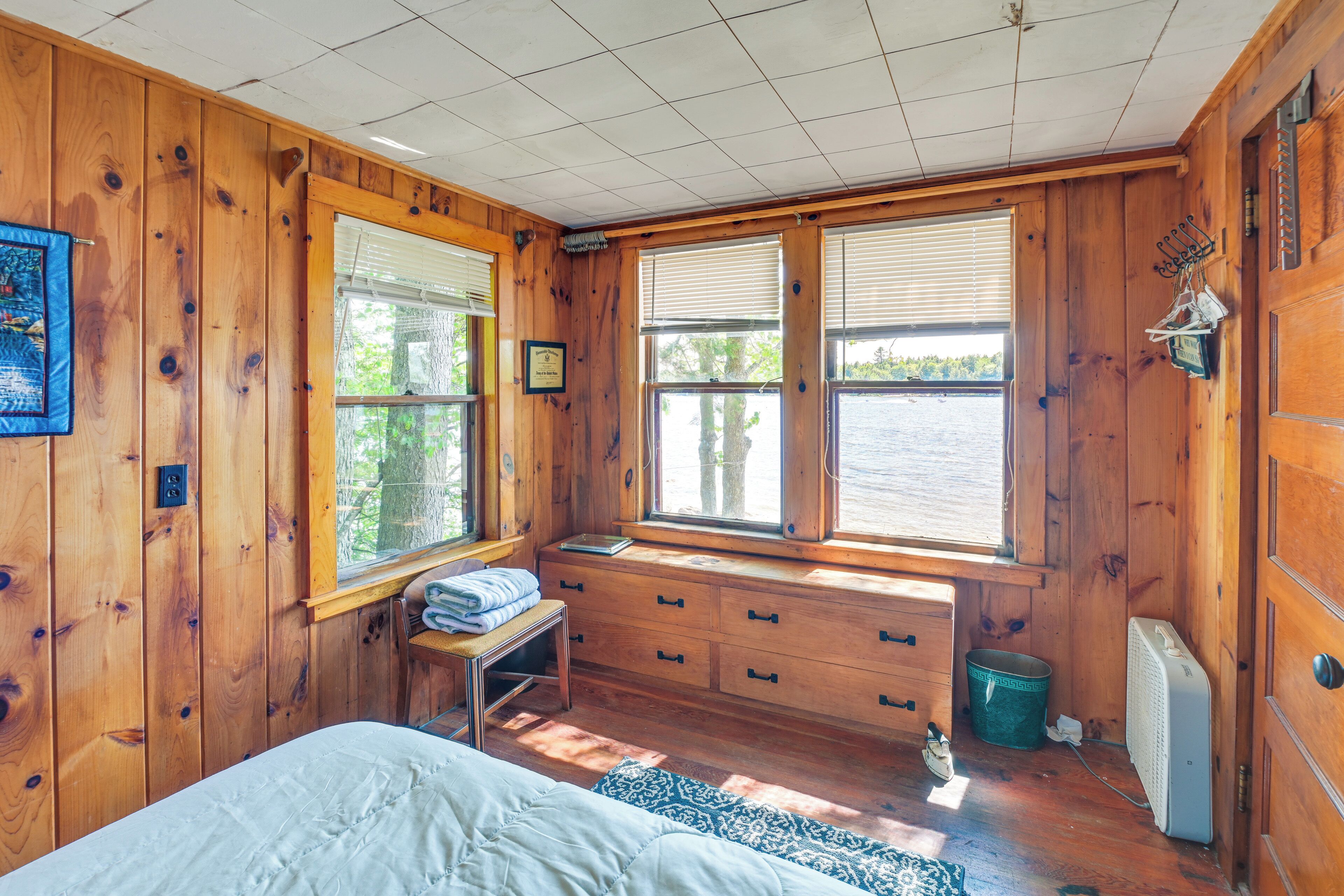 A wooden cabin bedroom with a bed, a bench, a dresser, and a window overlooking a body of water.