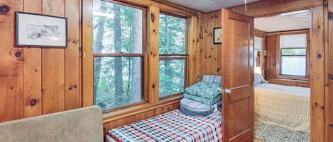 A cozy cabin room with a bed, a bench with a blanket, and large windows offering a view of the forest.