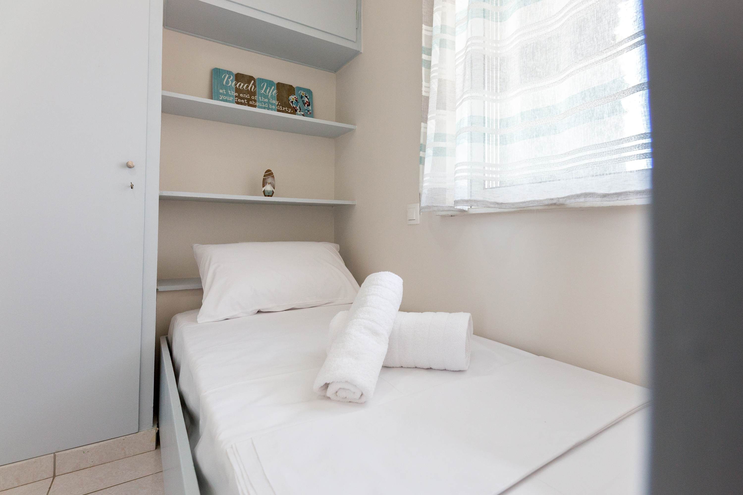 A neatly made bed with white linens and a rolled towel, a shelf with decorative items, and a window with sheer curtains.