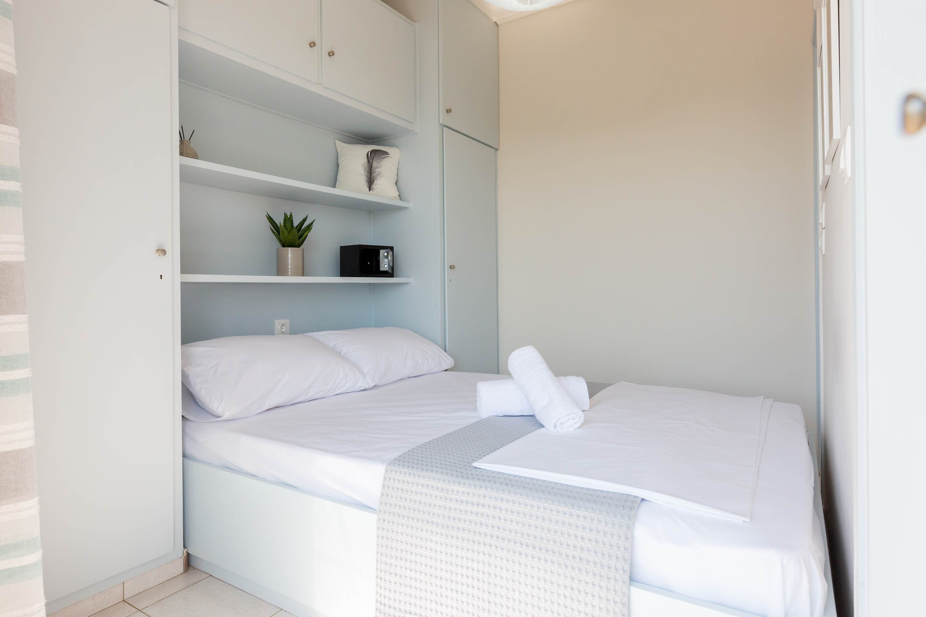 A neatly made bed with white linens and a rolled towel, a small potted plant on a shelf, and a black radio on another shelf.