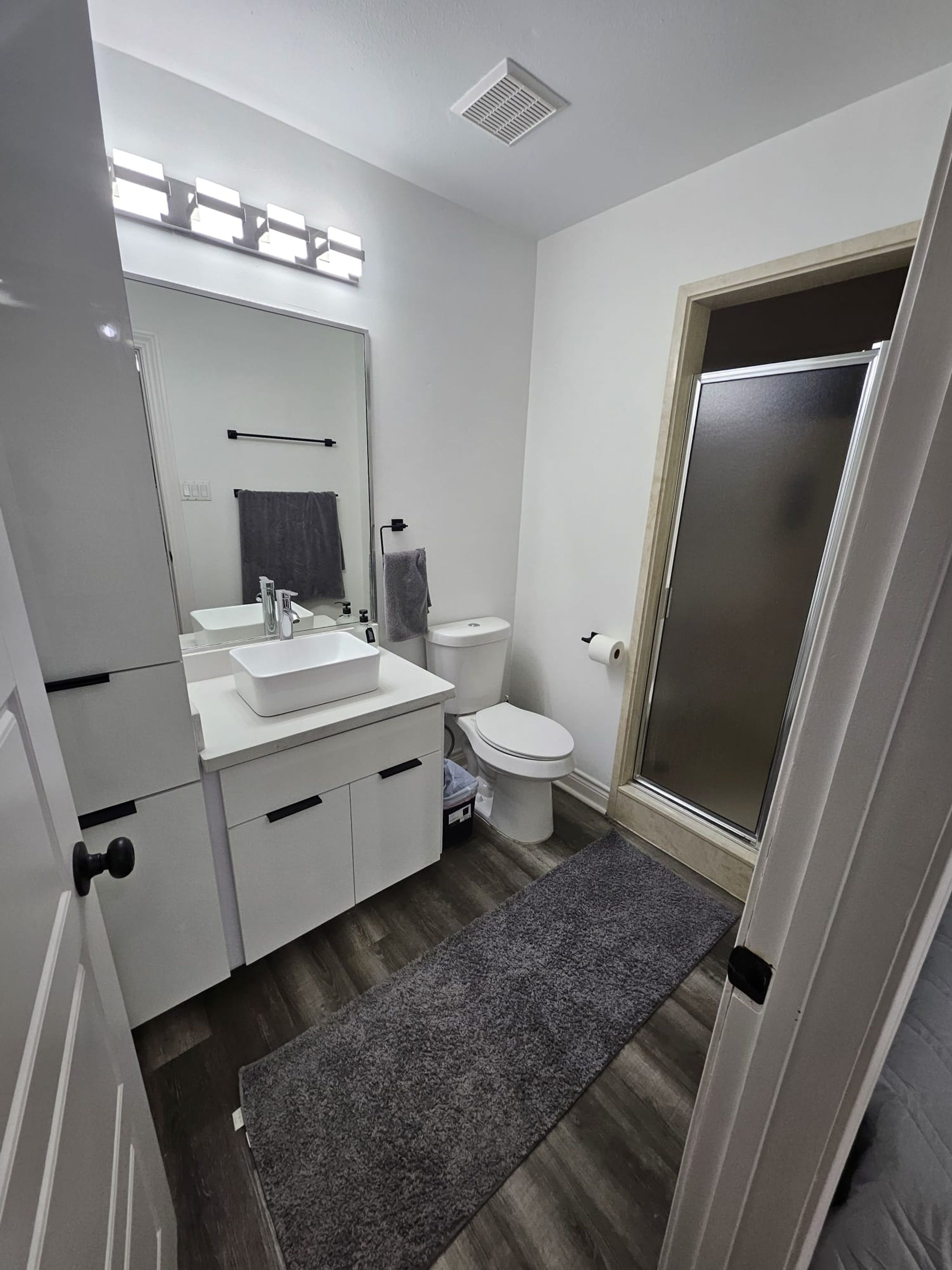 A modern bathroom with a white sink, mirror, and toilet. There is a towel rack above the sink and a small trash bin next to the toilet.