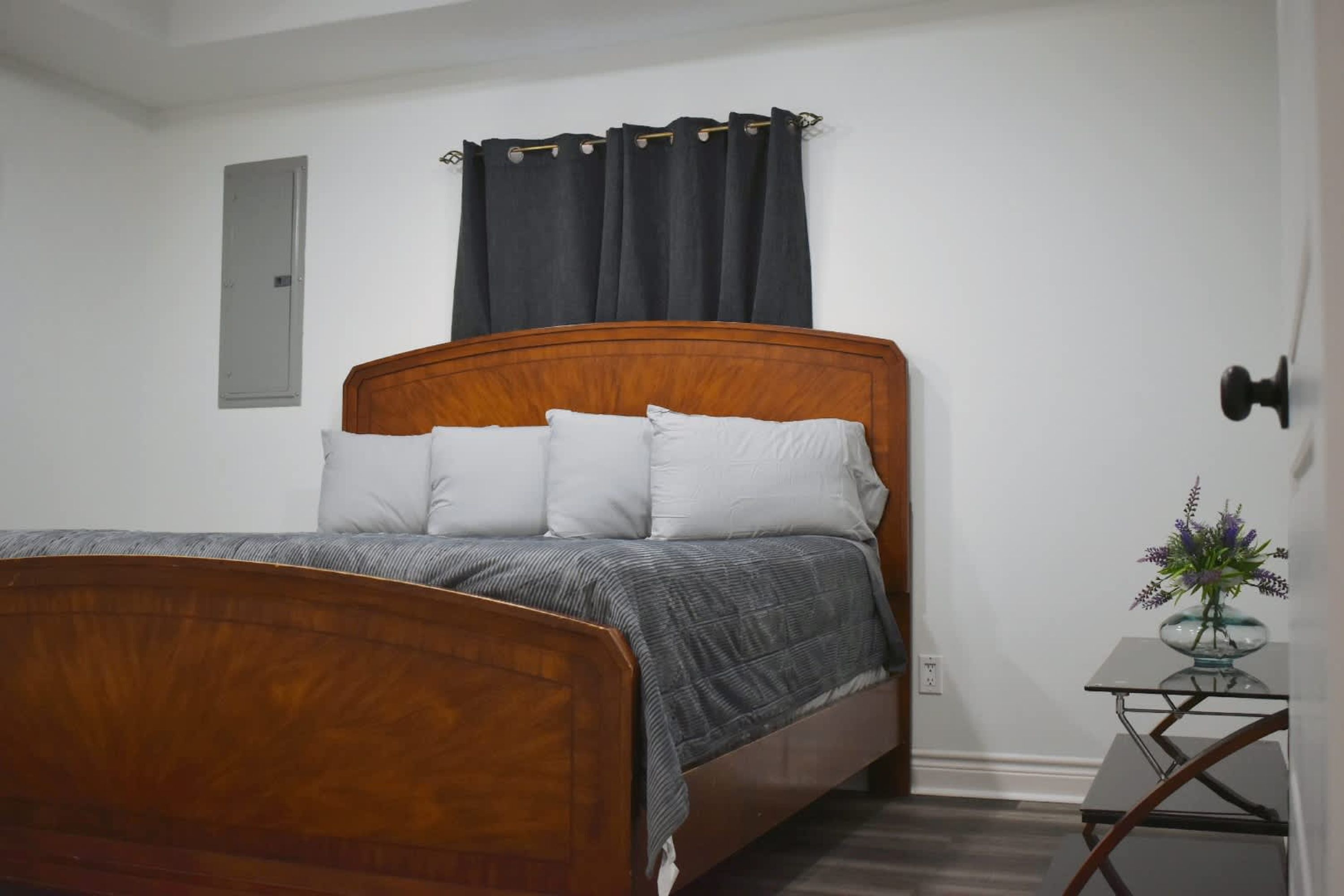 A bedroom with a wooden bed, white pillows, a gray blanket, a dark curtain, a small table with a vase, and a light switch on the wall.