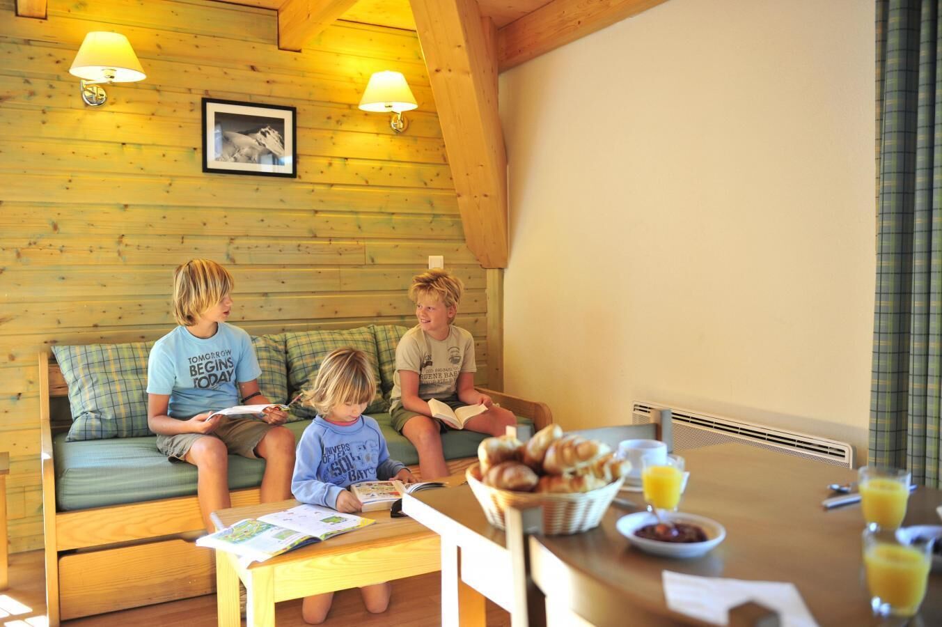 A cozy cabin interior with wooden walls, a sofa, a table with breakfast items, and two wall-mounted lamps.
