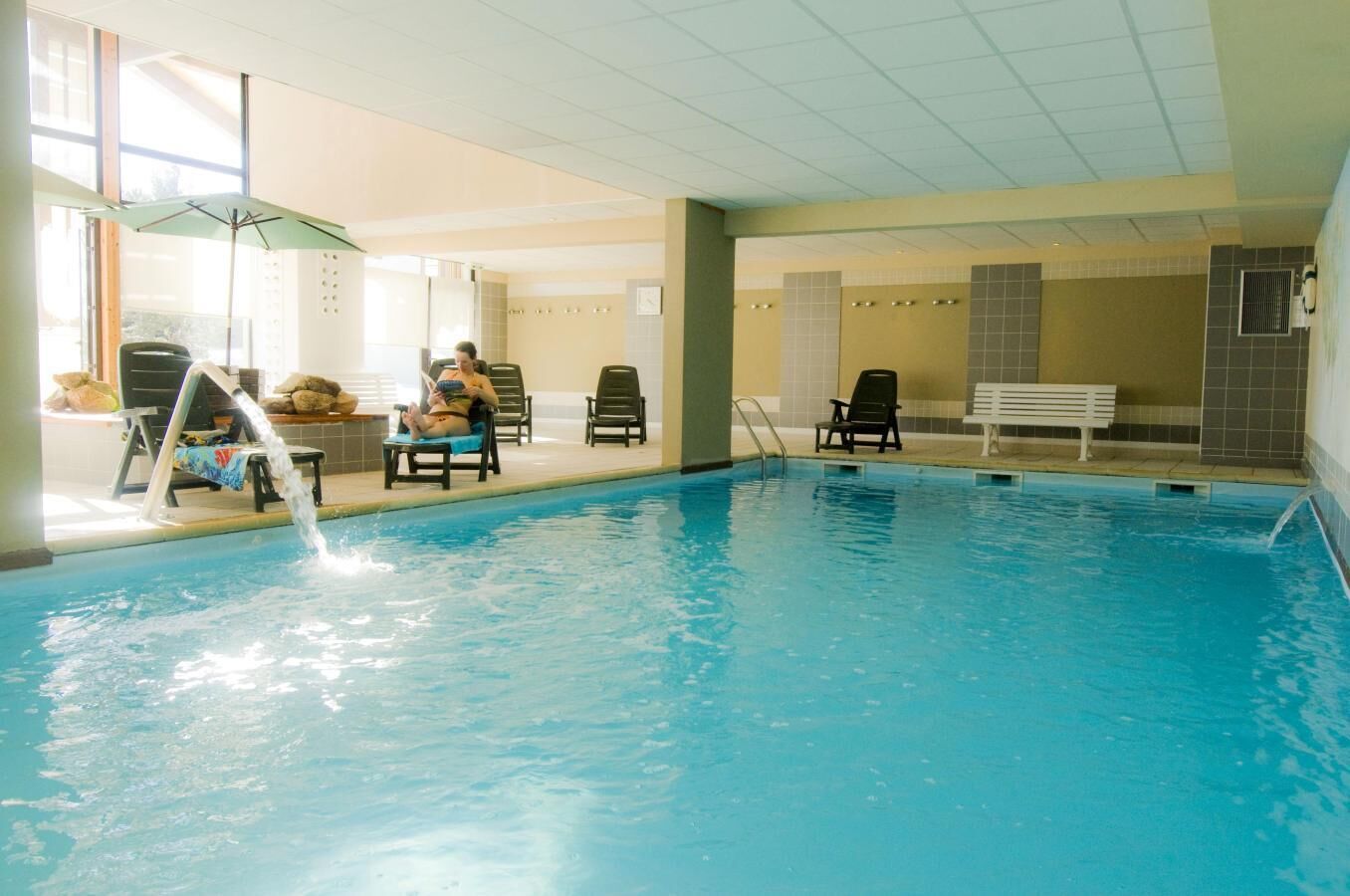 An indoor swimming pool with a water fountain, lounge chairs, and a bench.