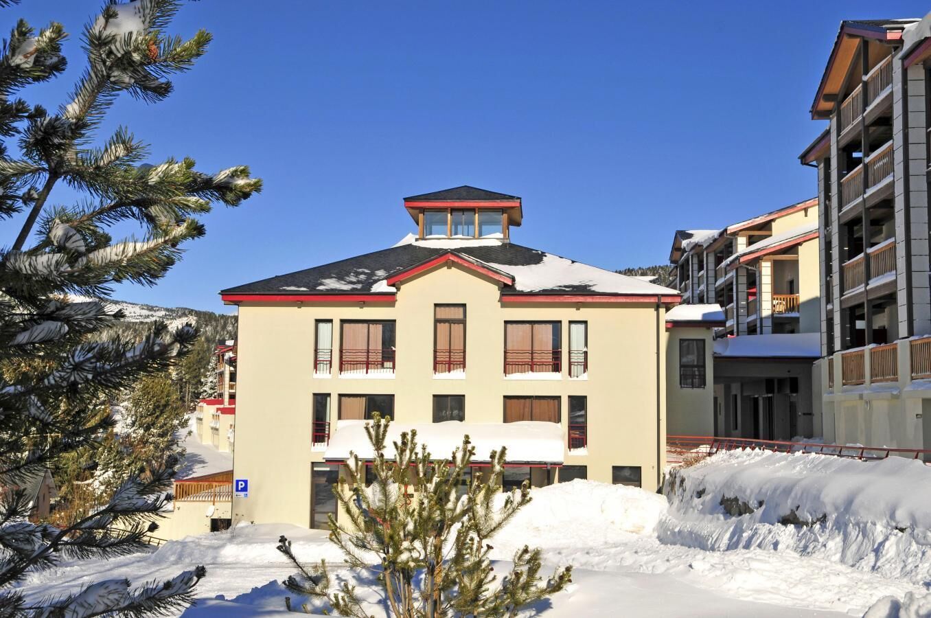 Un hotel de varios pisos con techo nevado y balcones, rodeado de nieve y pinos.