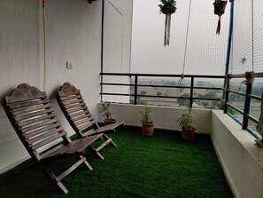 A balcony with two wooden chairs, a small artificial green turf, and three potted plants. - Anand's Aashiyana Homestay (Agra)