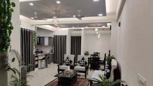 A modern living room with a dining area, featuring a large window, ceiling fans, and a kitchen in the background. - Anand's Aashiyana Homestay (Agra)