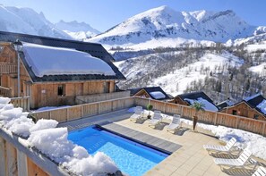 Un chalet en bois doté d’une piscine et de chaises longues, entouré de montagnes enneigées.