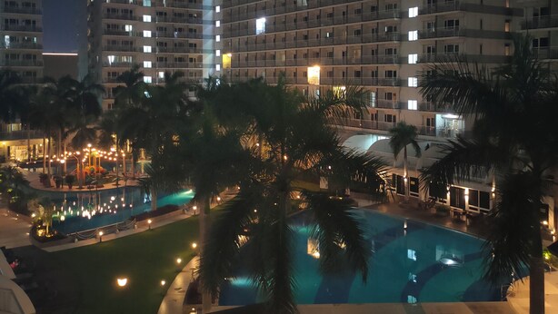 A nighttime view of a hotel with multiple high-rise buildings, a swimming pool, and surrounding palm trees. - 5-Minutes Walk to Mall of Asia, One-bedroom with Balcony at Shell Residences (Pasay)