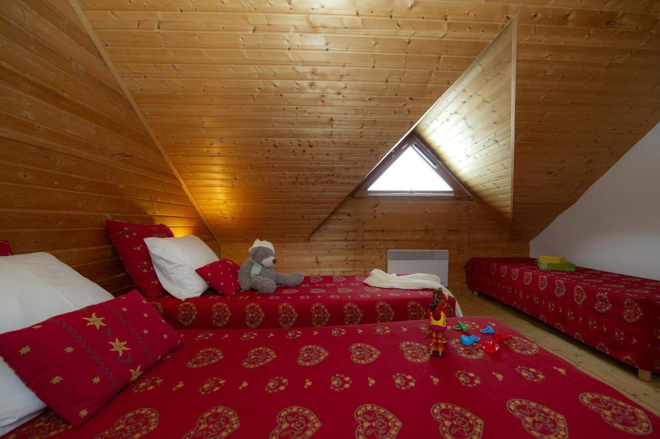 Une chambre mansardée chaleureuse avec des lambris en bois au plafond, deux lits avec une literie rouge et un puits de lumière.