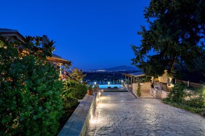 Un chemin de pierre mène à un espace piscine, d’où l’on peut admirer une montagne et les lumières de la ville en arrière-plan.
