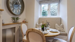 A dining area with a round table, chairs, a bench, a decorative clock, and a window with a view of greenery. - Antelope Cottage - Northleach, Cotswolds - near pubs (Northleach)