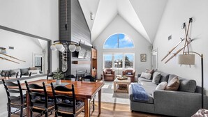 A spacious loft with a high ceiling, a dining area with wooden furniture, a gray sofa, and skis on the wall. - Apres Chalet: Winterplace Ski-in/Out pool+hot tubs (Ludlow)