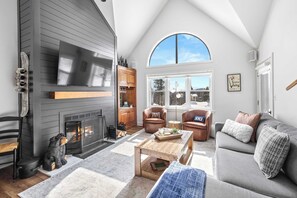 A cozy living room with a fireplace, leather chairs, a sofa, and a large window offering a view of the outdoors. - Apres Chalet: Winterplace Ski-in/Out pool+hot tubs (Ludlow)