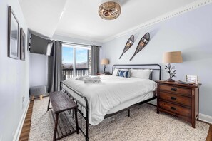A bedroom with a large bed, a bedside table, a bench, a TV mounted on the wall, and a window with curtains. - Apres Chalet: Winterplace Ski-in/Out pool+hot tubs (Ludlow)