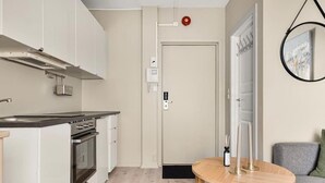 A compact kitchen with a stove, oven, and white cabinets. There's a round wooden table with candles and a gray sofa in the living area. - Cozy Studio in Bergen Center (Bergen)