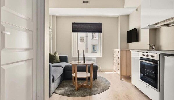 A compact kitchen with a built-in oven, a dining area with a round table and two chairs, a grey sofa, and a window with a dark blind. - Cozy Studio in Bergen Center (Bergen)